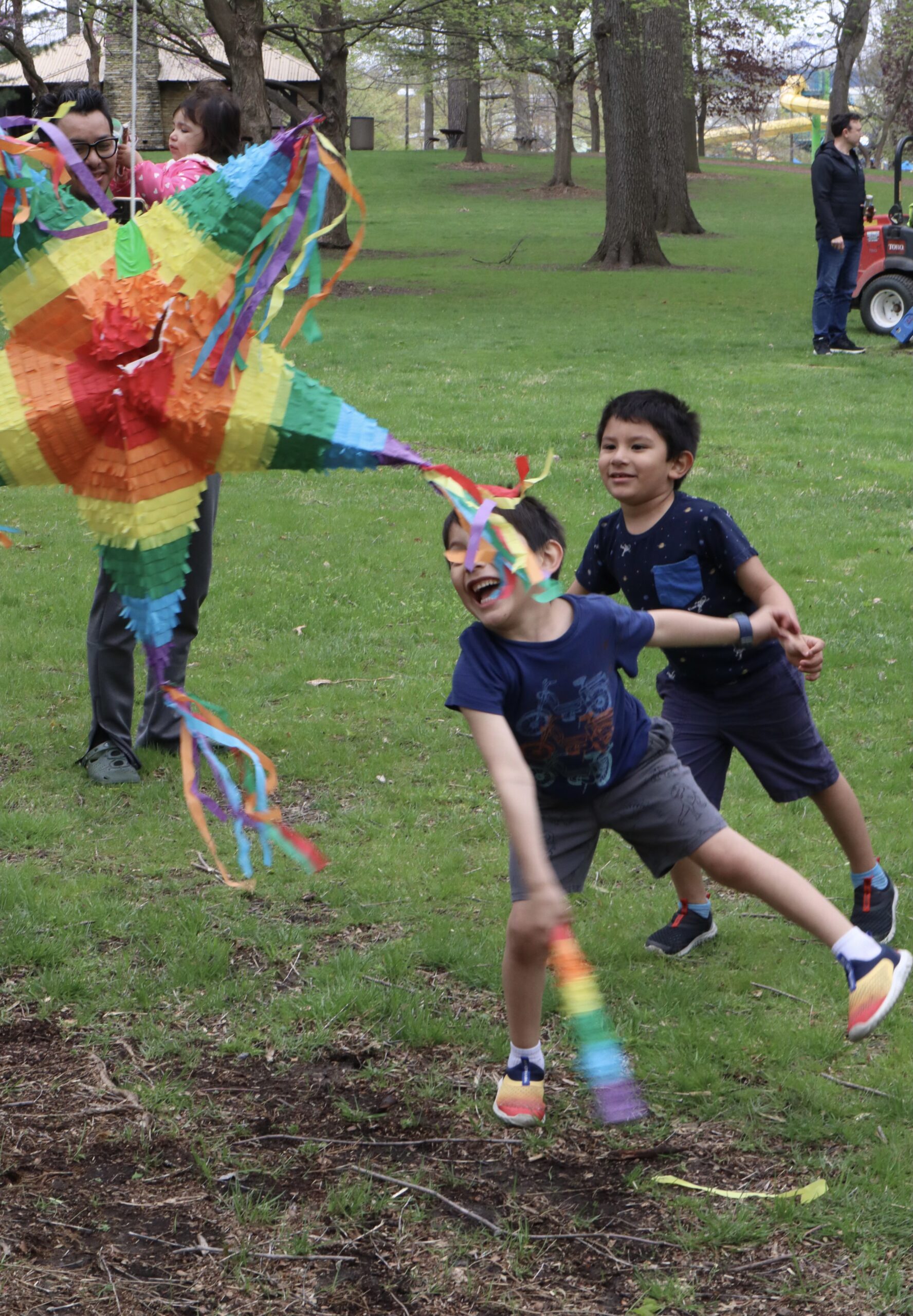 IMG_1232 Two young children are smiling, trying to swing at a pinata. A father and his daughter watch as the children are having fun.