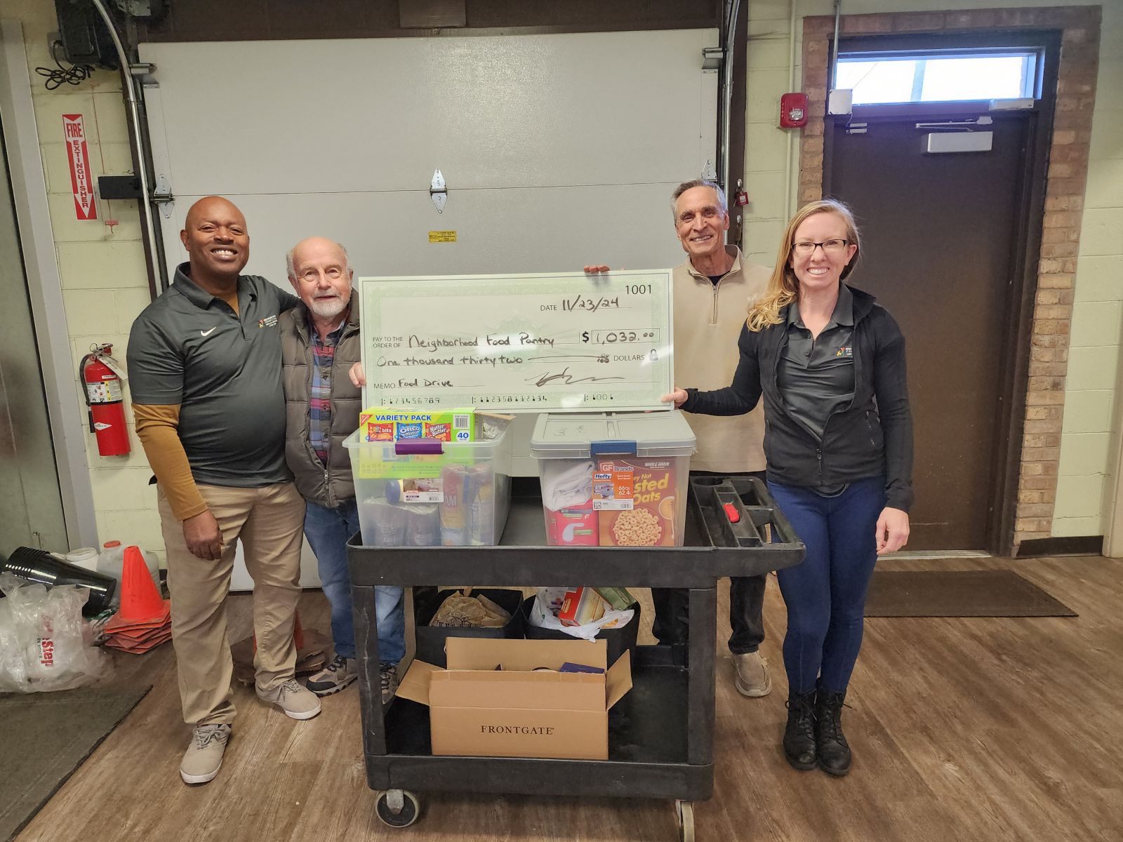 Four people surround a cart of food in a warehouse. The are holding a large check made out to Neighborhood Food Pantry in the amount of $1,032