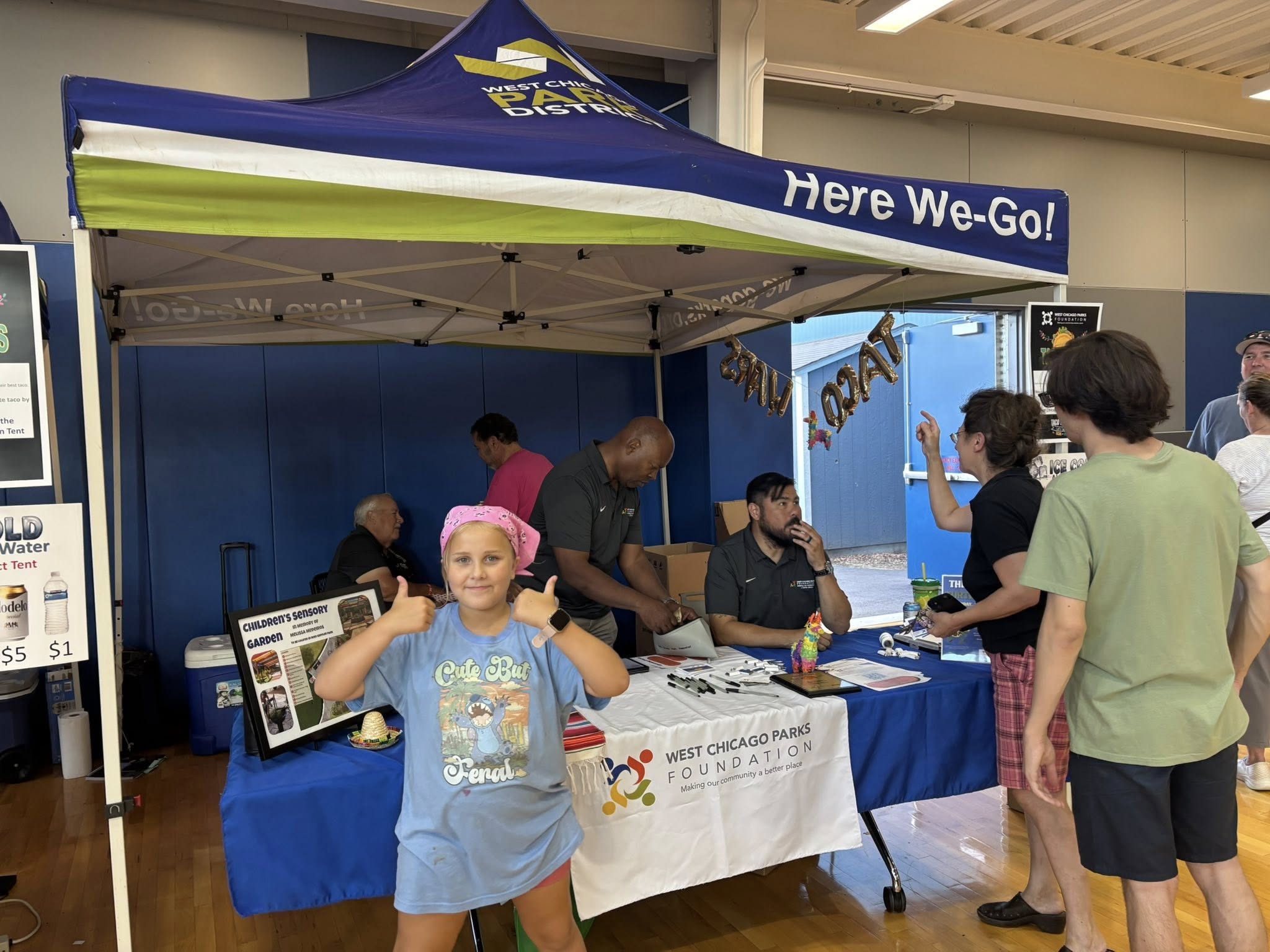 tw2 members of the west chicago parks foundation sell tickets under a canopy for taco wars 2025, while a small child gives the thumbs up in front of the table
