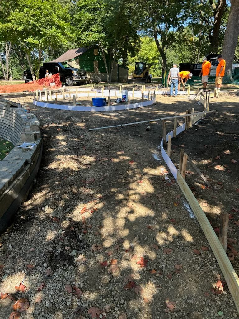 Construction in a park setting, showing workers and heavy equipment in the background. In the foreground, a curved section of concrete formwork made of white material and wooden stakes follows a path of dirt and gravel, leading toward a circular area. A low stone wall is visible on the left.