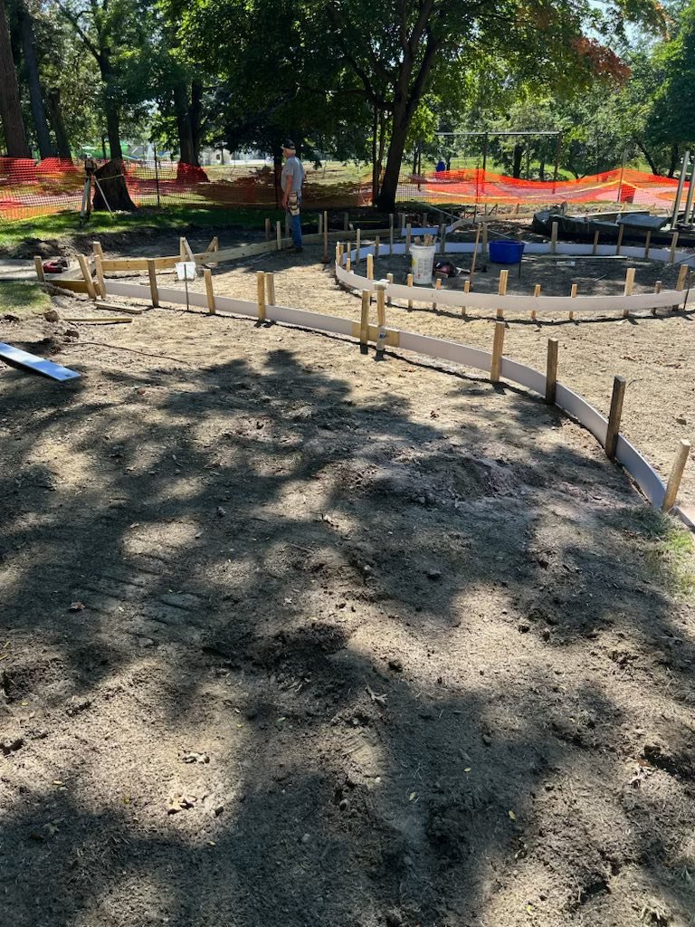 A close-up view of the initial construction phase on a path. Wooden stakes and white, flexible material are used to create the formwork for a curved pathway. A worker is visible in the middle background near more complex, raised formwork.