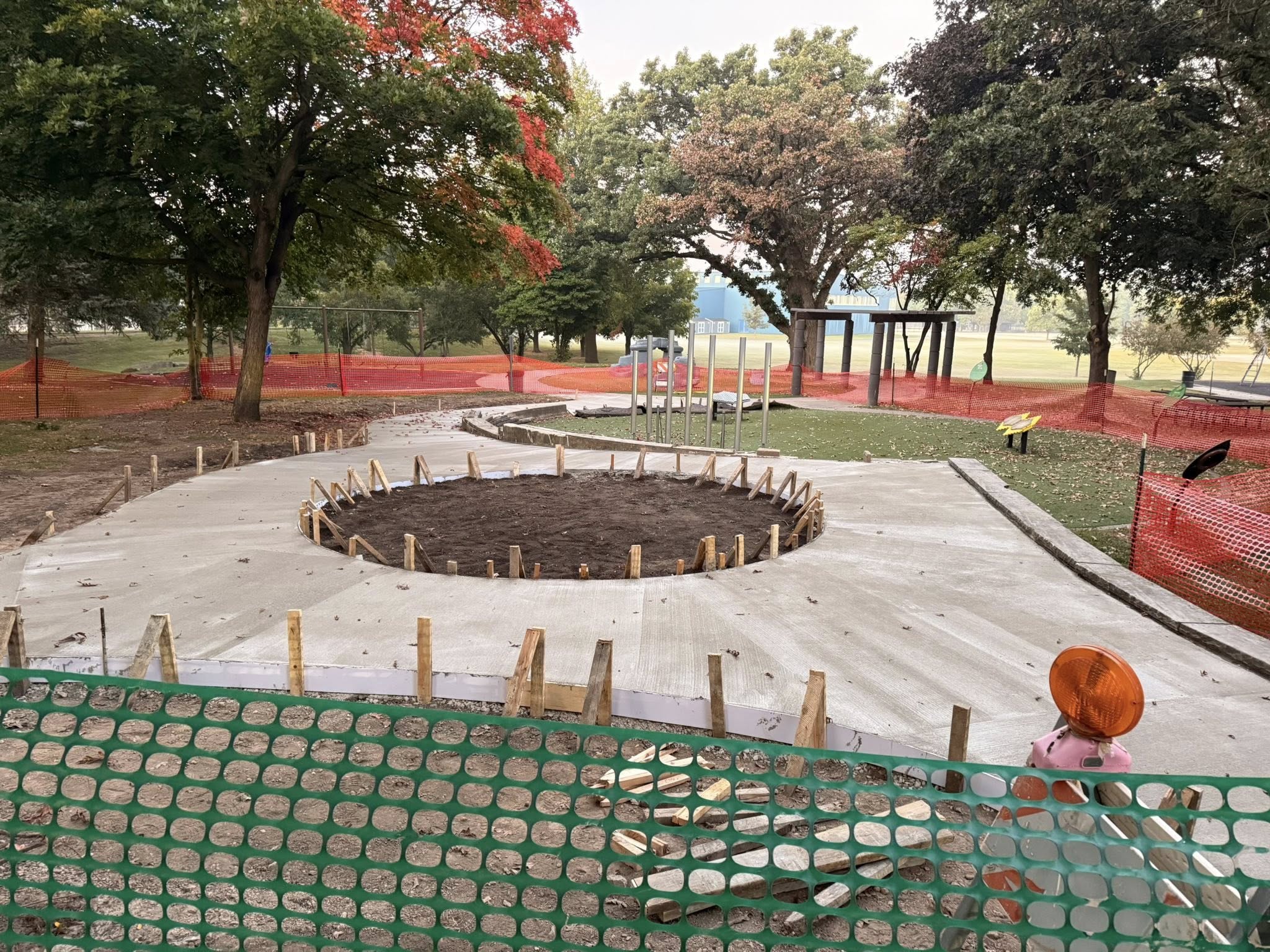 The most complete phase of construction shown. A large concrete slab has been poured and cured, featuring a circular area of exposed dirt in the center for planting. Wooden stakes form the edge of the central planting area. Orange and green safety fencing surrounds the construction site. A playground area is visible in the background among mature trees.